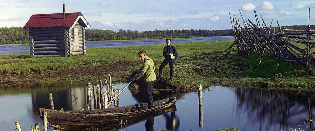 Two men and a boat with a little log building and lake in background