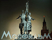 Statue of young man and woman holding the hammer and sickle aloft. Word Mosfilm is at their feet. Kremlin clock tower is behind them to one side.